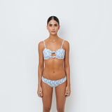 A woman standing against a white background, wearing a blue geometric patterned bikini top with peekaboo detail and matching bikini bottoms.