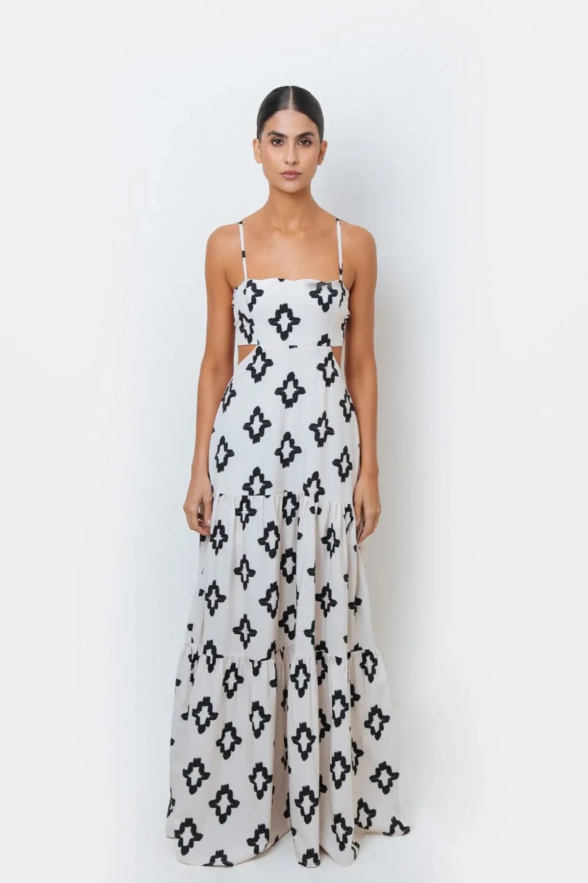 A woman modeling a sleeveless, spaghetti-strap maxi dress with a geometric black and white pattern, featuring cut-out side detail with adjustable back ties.