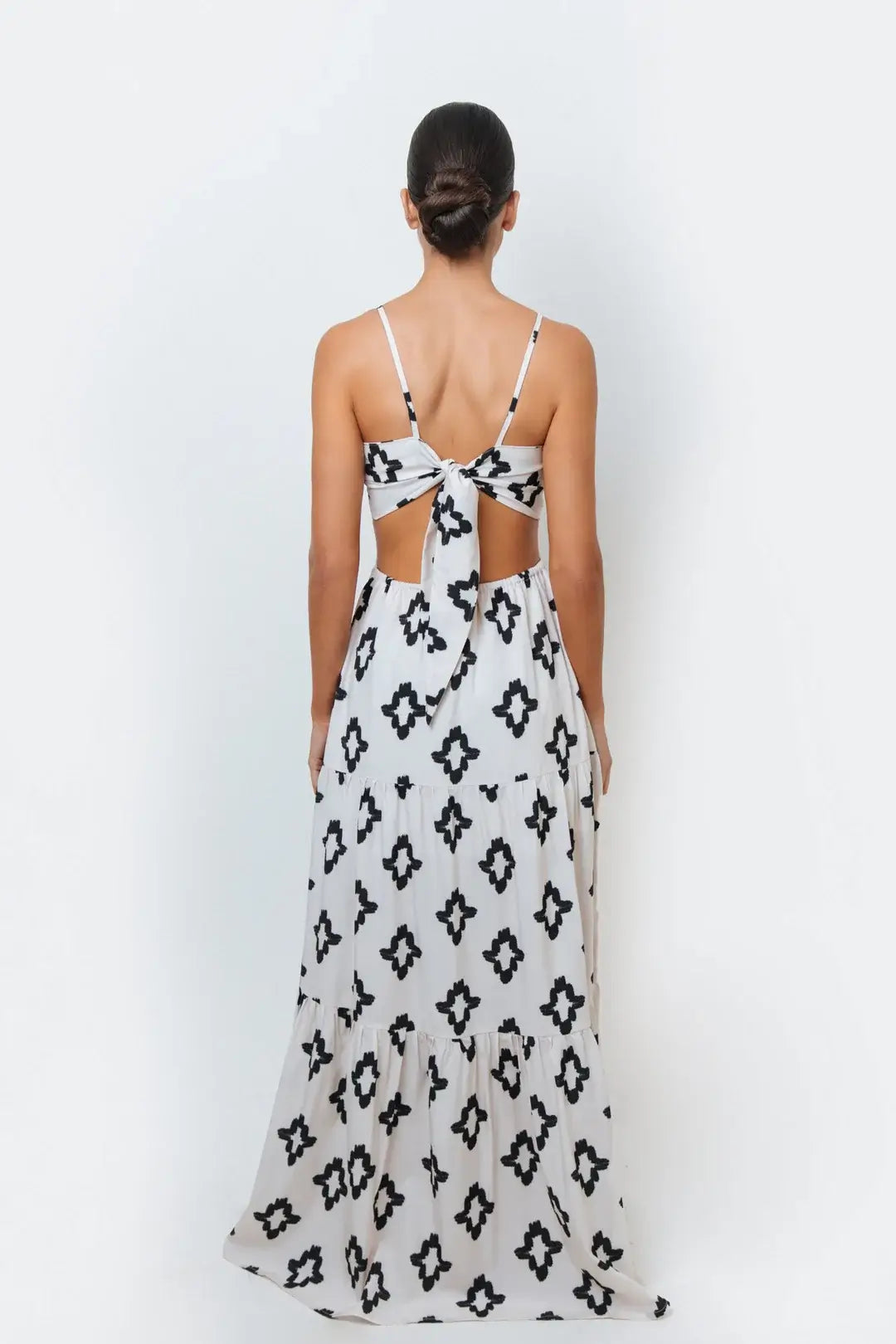A woman modeling a sleeveless, spaghetti-strap maxi dress with a geometric black and white pattern, featuring cut-out side detail with an adjustable back-tie.