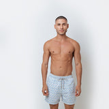 A man standing against a white background, wearing blue and white geometric patterned swim trunks.