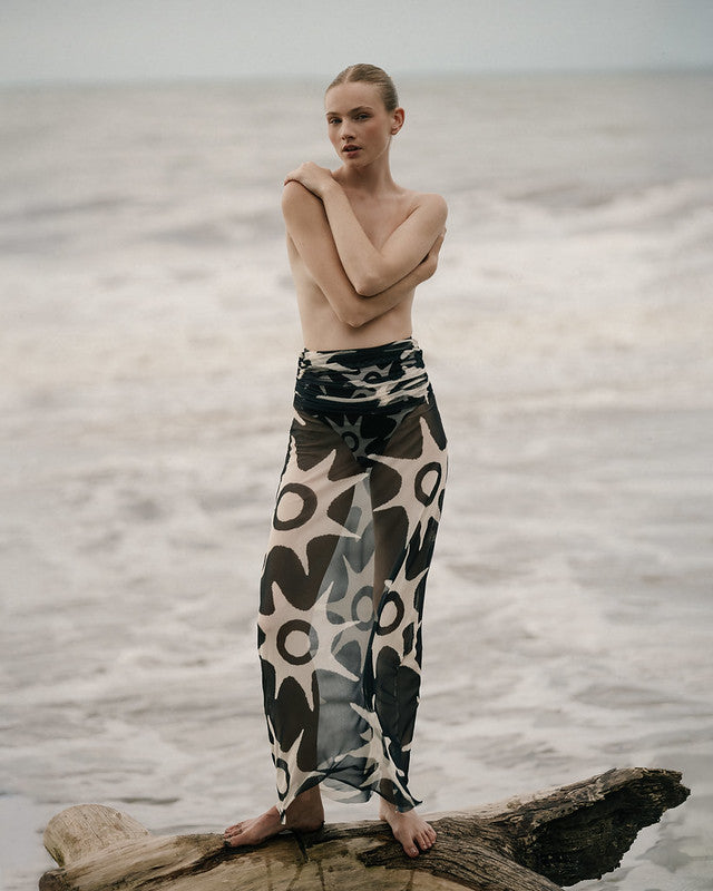 Woman standing on a log by a body of water wearing a patterned skirt.