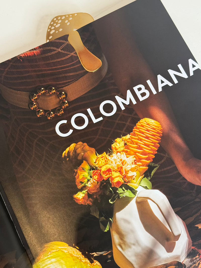 Close up image of a mushroom-shaped bookmark in gold, on a book called, "Colombiana." The purpose of this image is to highlight the bookmark, which is handmade by PAJAROLIMON.