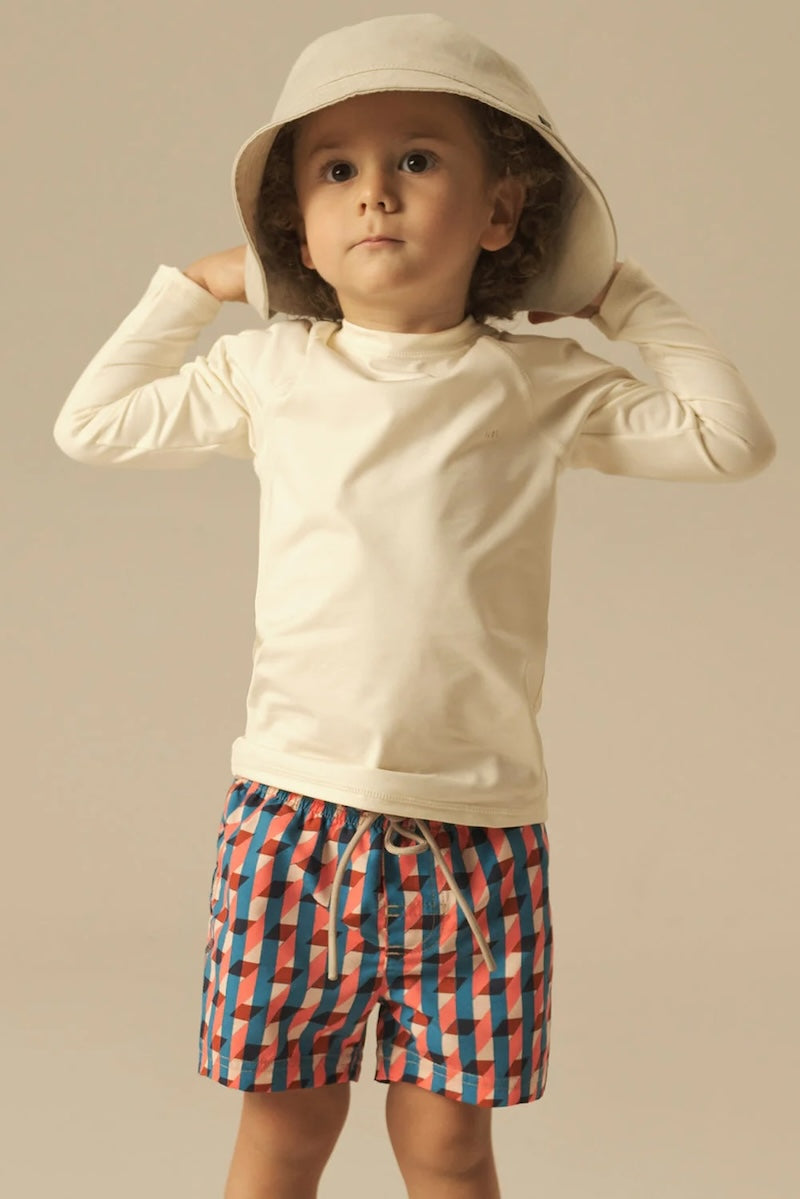 Child wearing a beige long-sleeve shirt and colorful geometric swim shorts against a neutral background.
