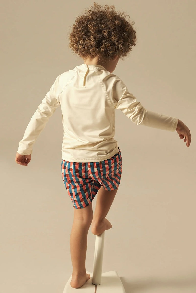 Child wearing a cream long-sleeve shirt and pink, red and blue checkered shorts on a beige background.