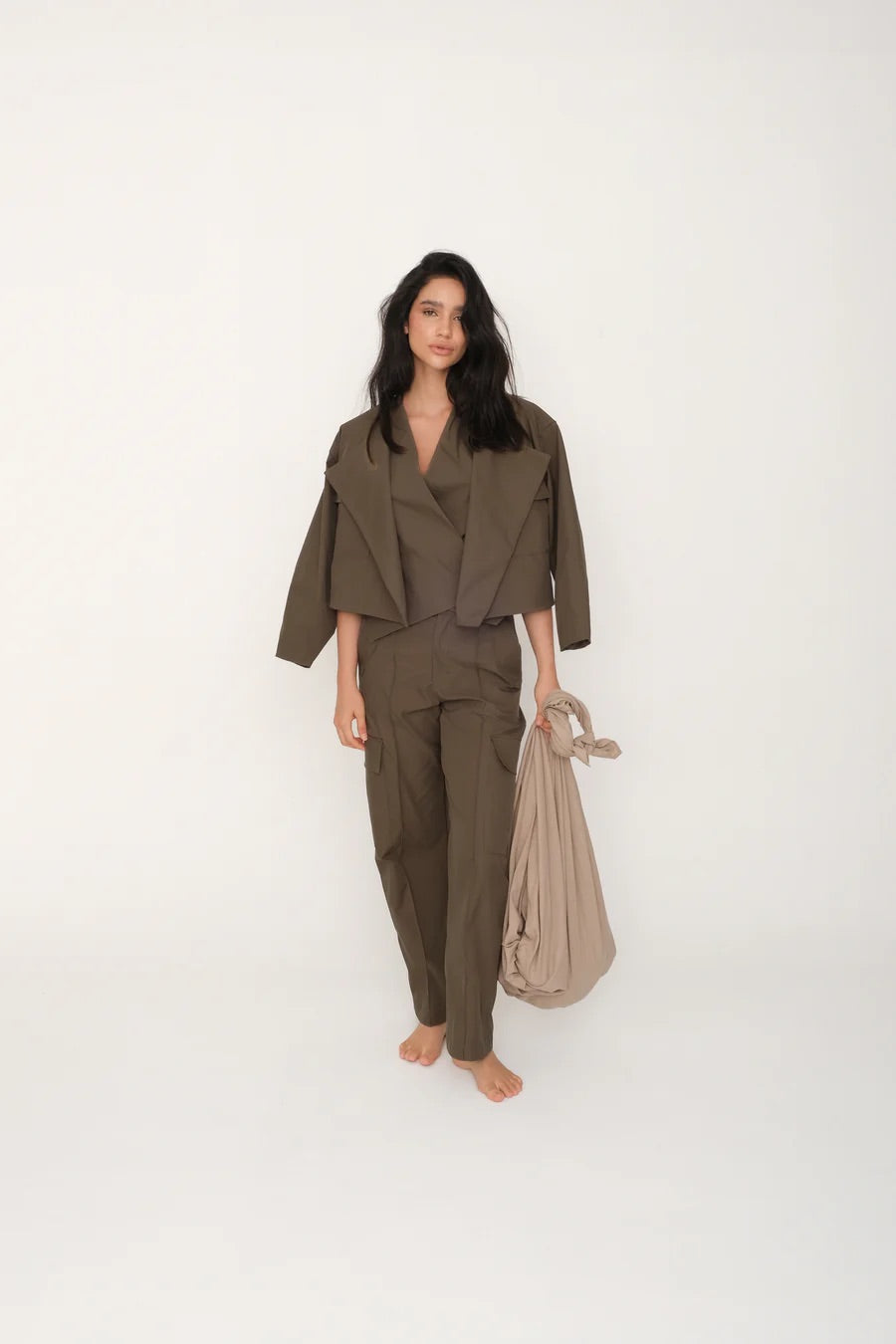 Woman wearing an olive green top and pants set with a matching jacket holding a beige bag on a white background.