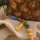 A gold-plated pineapple pendent necklace with oversized multicolored beads displayed in front of a pineapple fruit.