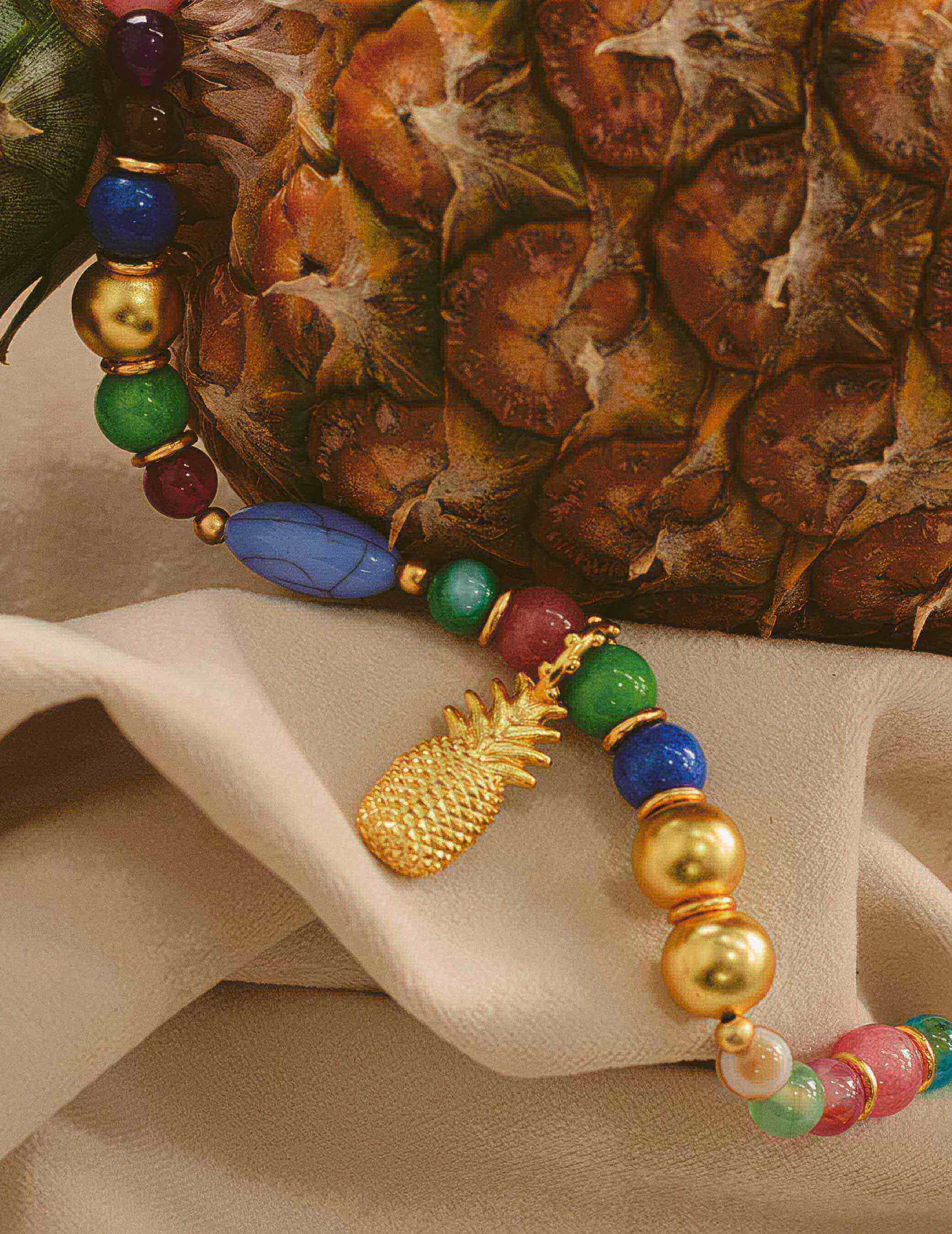 A gold-plated pineapple pendent necklace with oversized multicolored beads displayed in front of a pineapple fruit.