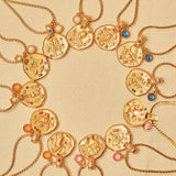 A collection of 24k gold-plated necklaces with different zodiac sign pendants, each accompanied by a natural stone charm, displayed on a beige background.