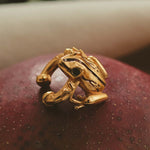 A gold-plated earcuff of a Coqui frog, displayed on a red surface.