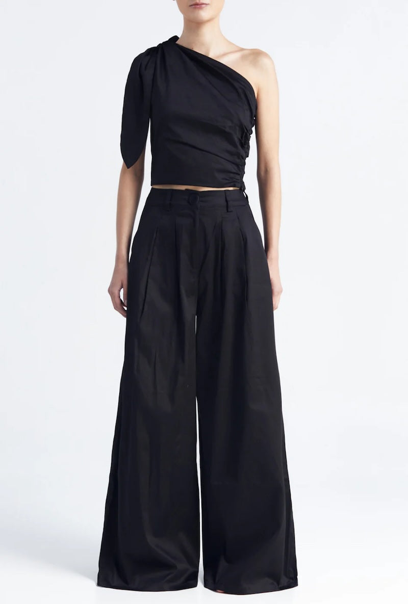 Image of a woman wearing a one shoulder top with matching pants in black. The image is set against a white background. The purpose of this image is to highlight the clothing by Encantadore because it's for sale at EVAMAIA boutique in Studio City, CA.