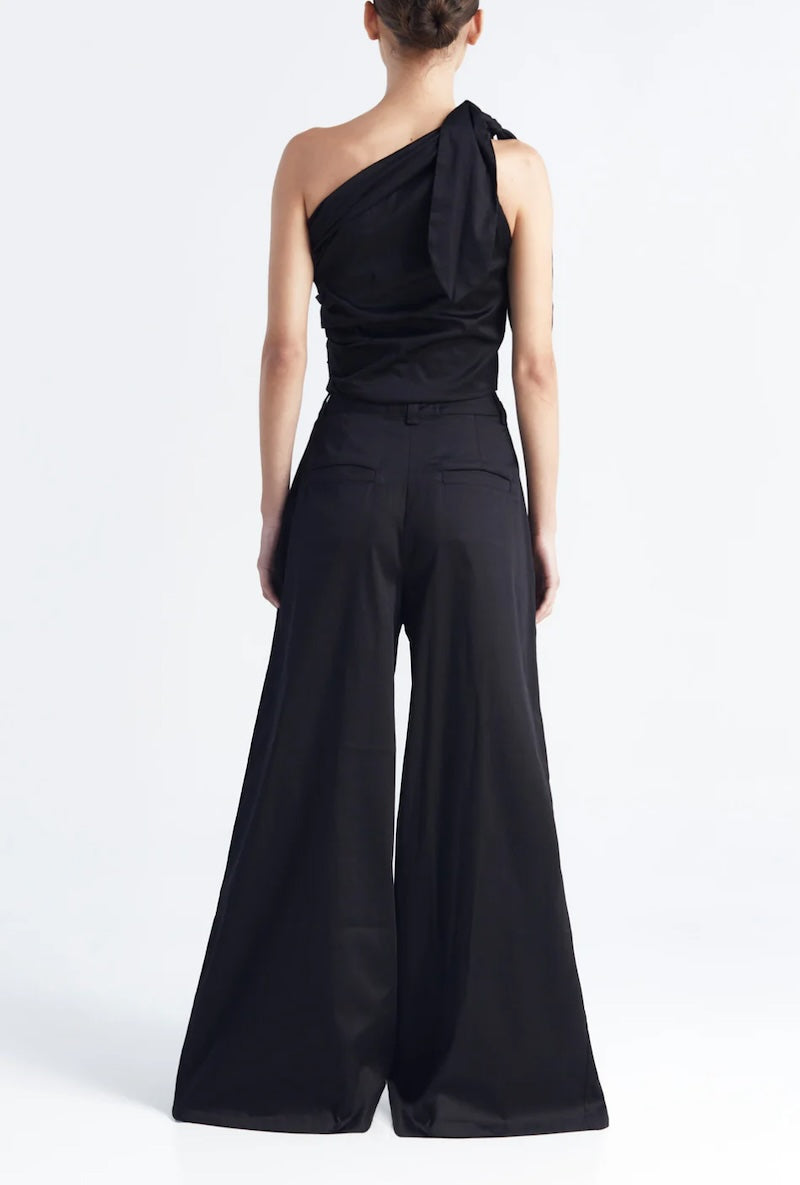 Image of a woman wearing a one shoulder top with matching pants in black. The image is set against a white background. The purpose of this image is to highlight the clothing by Encantadore because it's for sale at EVAMAIA boutique in Studio City, CA.
