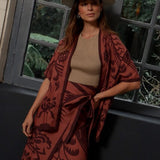 Woman in beige hat and patterned burnt orange and burgundy kimono standing by a window.