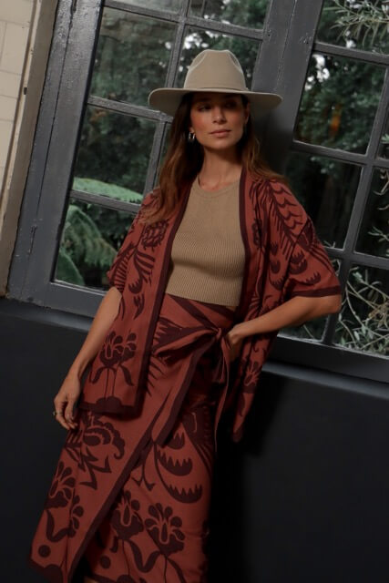 Woman in beige hat and patterned burnt orange and burgundy kimono standing by a window.