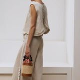Woman in a light-colored outfit holding a woven basket style purse, standing against a white wall.