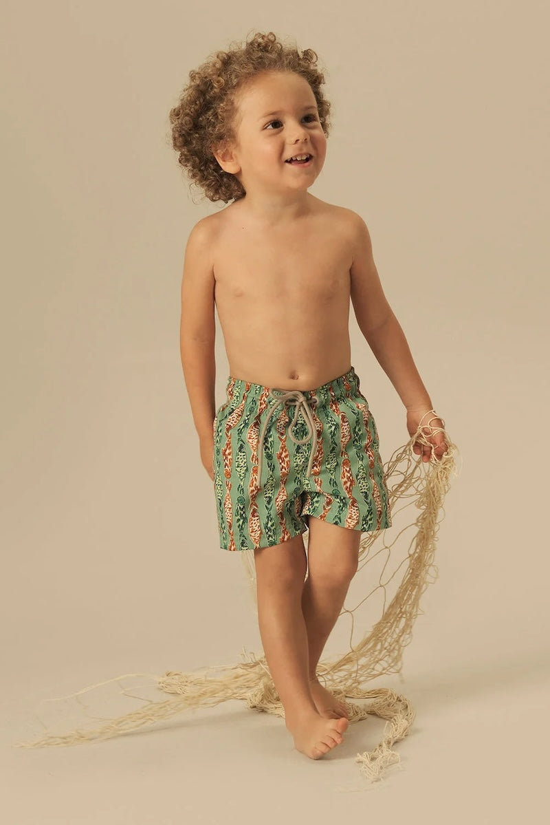 Child wearing green and orange fish patterned shorts standing on a beige background.