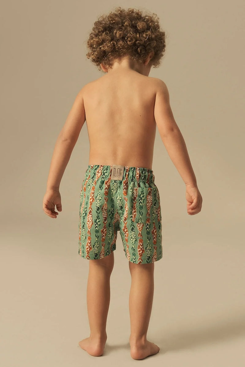 Child wearing green and orange fish patterned swim shorts on a beige background.