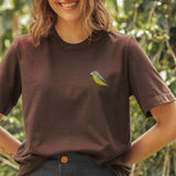 Woman wearing a brown t-shirt with a colorful bird design, standing outdoors with greenery in the background.