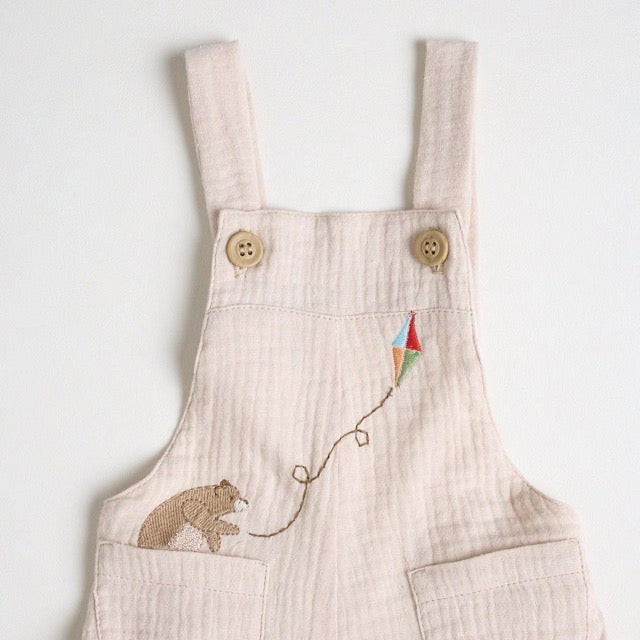 A close-up a beige short-sleeved overall with bear embroidery on the front, standing against a white background.