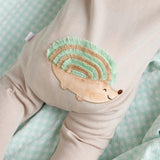 A close-up of a beige short-sleeve cotton onesie with a hand-embroidered hedgehog on the chest and a matching applique on the bottom, lying on a similar colored fabric.