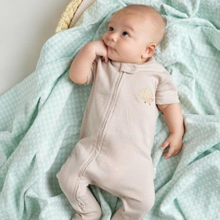 A baby wearing a beige short-sleeve cotton onesie with a hand-embroidered animal on the chest and a matching applique on the bottom, lying on a similar colored fabric.