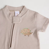 A close-up of a beige short-sleeve cotton onesie with a hand-embroidered hedgehog on the chest and a matching applique on the bottom, lying on a similar colored fabric.