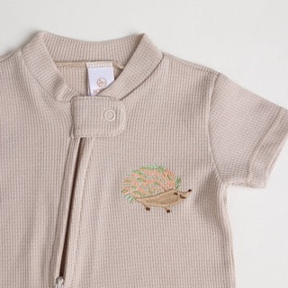 A close-up of a beige short-sleeve cotton onesie with a hand-embroidered hedgehog on the chest and a matching applique on the bottom, lying on a similar colored fabric.