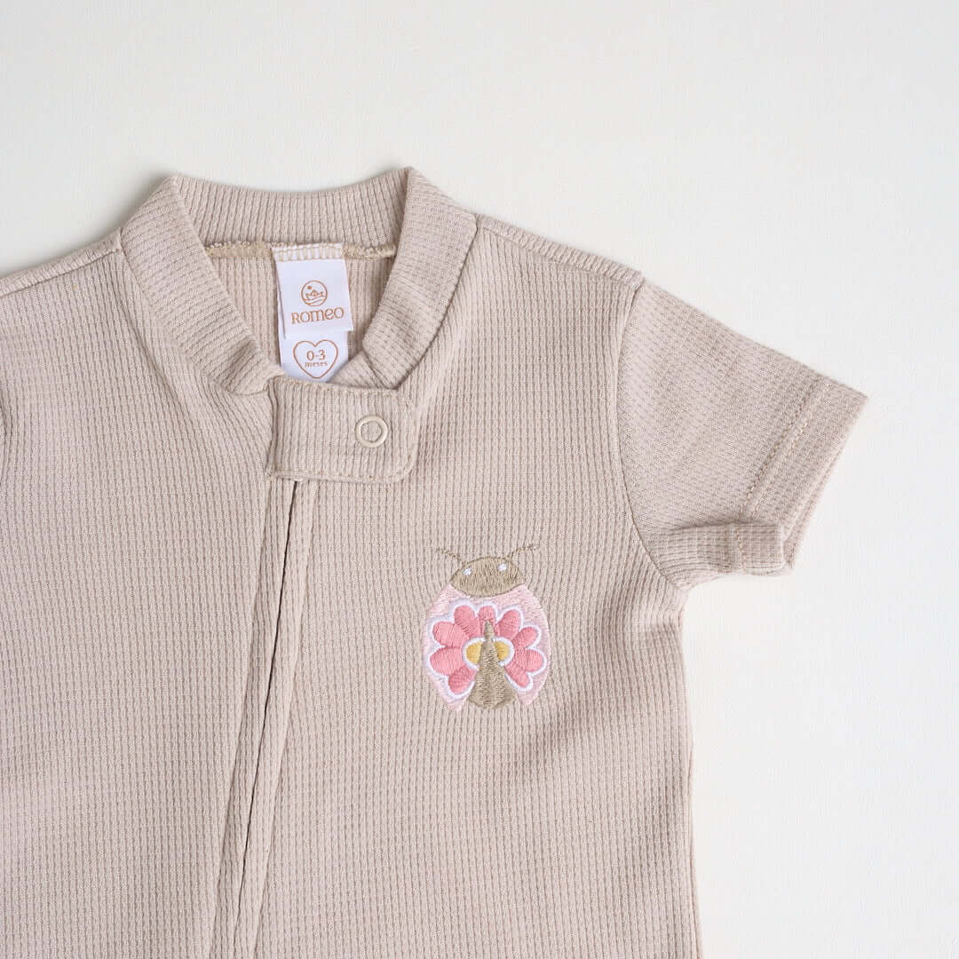Close-up of a beige short-sleeve cotton onesie with a hand-embroidered ladybug on the chest and a matching applique on the bottom.