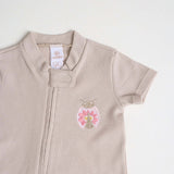 Close-up of a beige short-sleeve cotton onesie with a hand-embroidered ladybug on the chest and a matching applique on the bottom.