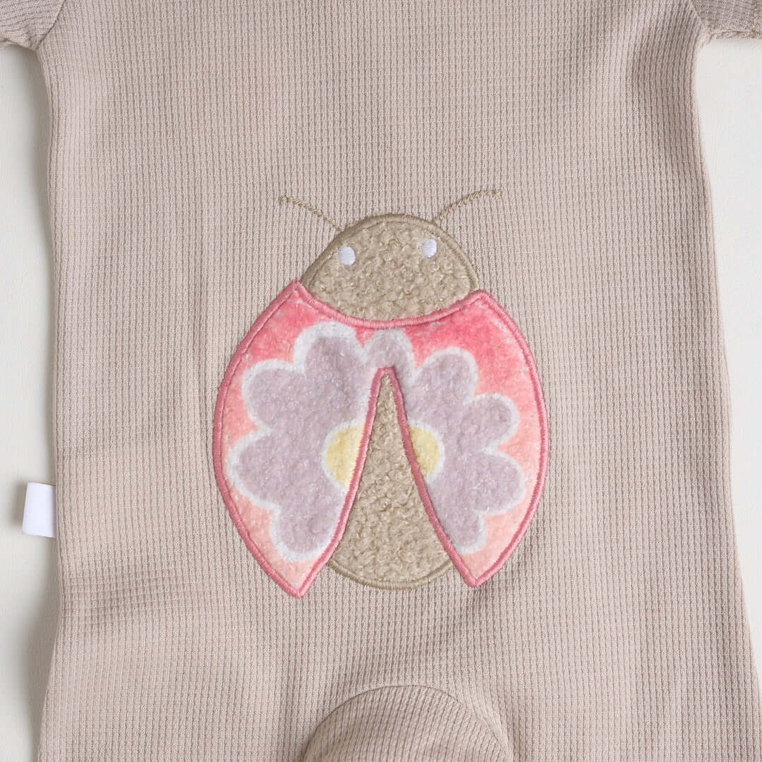 Close-up of a beige short-sleeve cotton onesie with a hand-embroidered ladybug on the chest and a matching applique on the bottom.