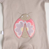 Close-up of a beige short-sleeve cotton onesie with a hand-embroidered ladybug on the chest and a matching applique on the bottom.
