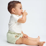 A baby wearing a white bodysuit and green diaper cover with wooden button accents and a embroidered bear detail.