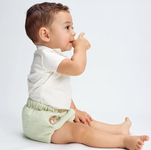 A baby wearing a white bodysuit and green diaper cover with wooden button accents and a embroidered bear detail.