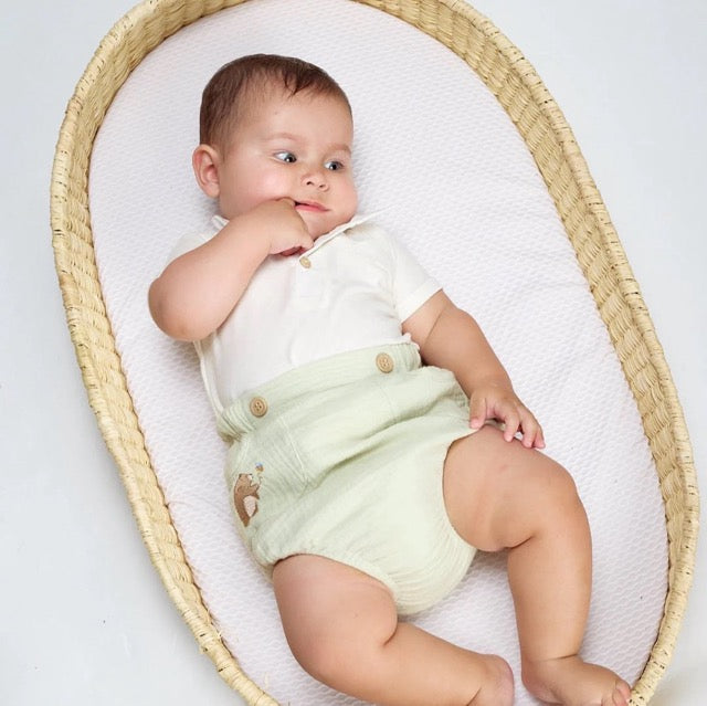 A baby wearing a white bodysuit and green diaper cover with wooden button accents and a embroidered bear detail.