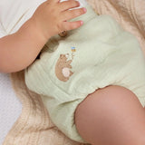 A close-up of a baby wearing a white bodysuit and green diaper cover with wooden button accents and a embroidered bear detail.