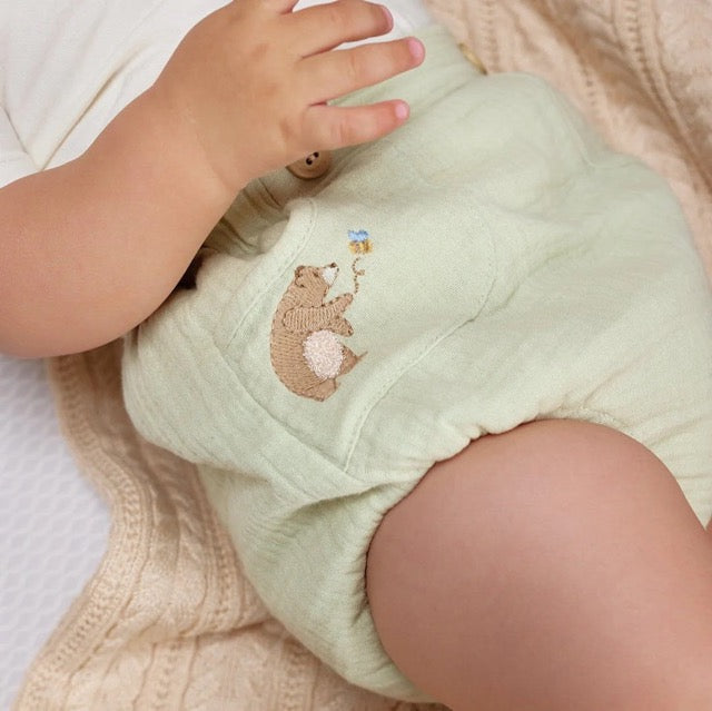 A close-up of a baby wearing a white bodysuit and green diaper cover with wooden button accents and a embroidered bear detail.