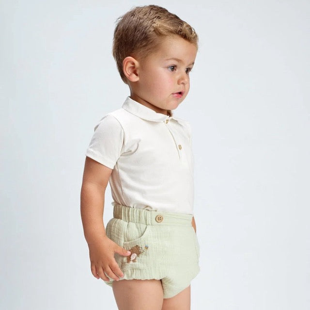 A baby wearing a white bodysuit and green diaper cover with wooden button accents and a embroidered bear detail.