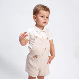 A toddler wearing a short-sleeved polo with a beige overall that has bear embroidery on the front, standing against a white background.