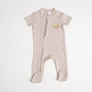 A close-up of a beige short-sleeve cotton onesie with a hand-embroidered hedgehog on the chest and a matching applique on the bottom, lying on a similar colored fabric.