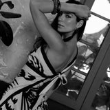 A black and white photo of a woman wearing a floral white and black sari styled as a wrap dress, standing indoors.