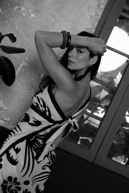 A black and white photo of a woman wearing a floral white and black sari styled as a wrap dress, standing indoors.