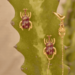 Gold beetle-shaped earrings with red gemstones on a cactus plant.