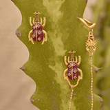 Gold beetle-shaped earrings with red gemstones on a cactus plant.