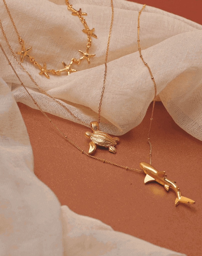 Gold necklaces with starfish, turtle and whale pendants on a beige fabric background.
