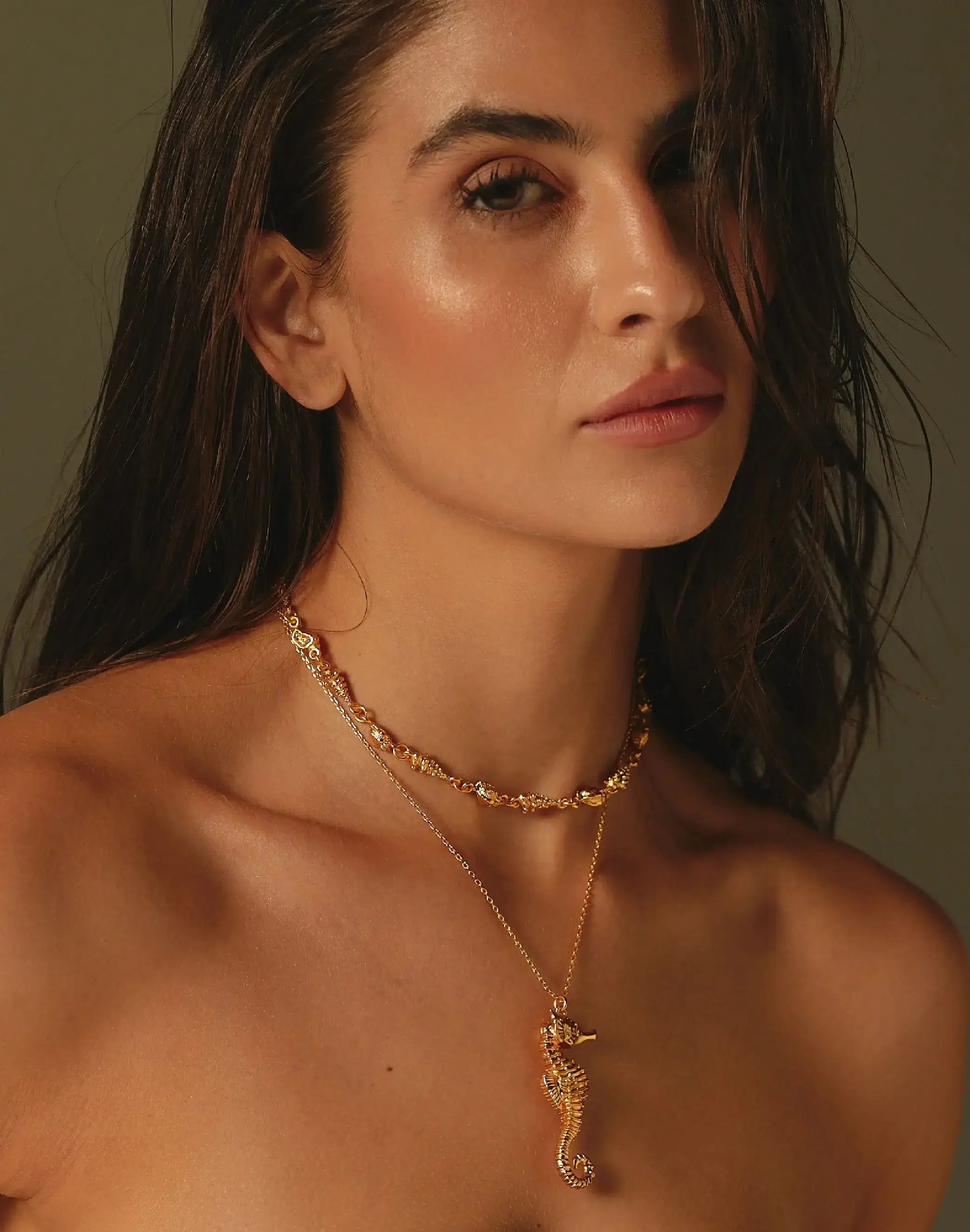 Woman wearing a gold necklace with a seahorse pendant against a neutral background.