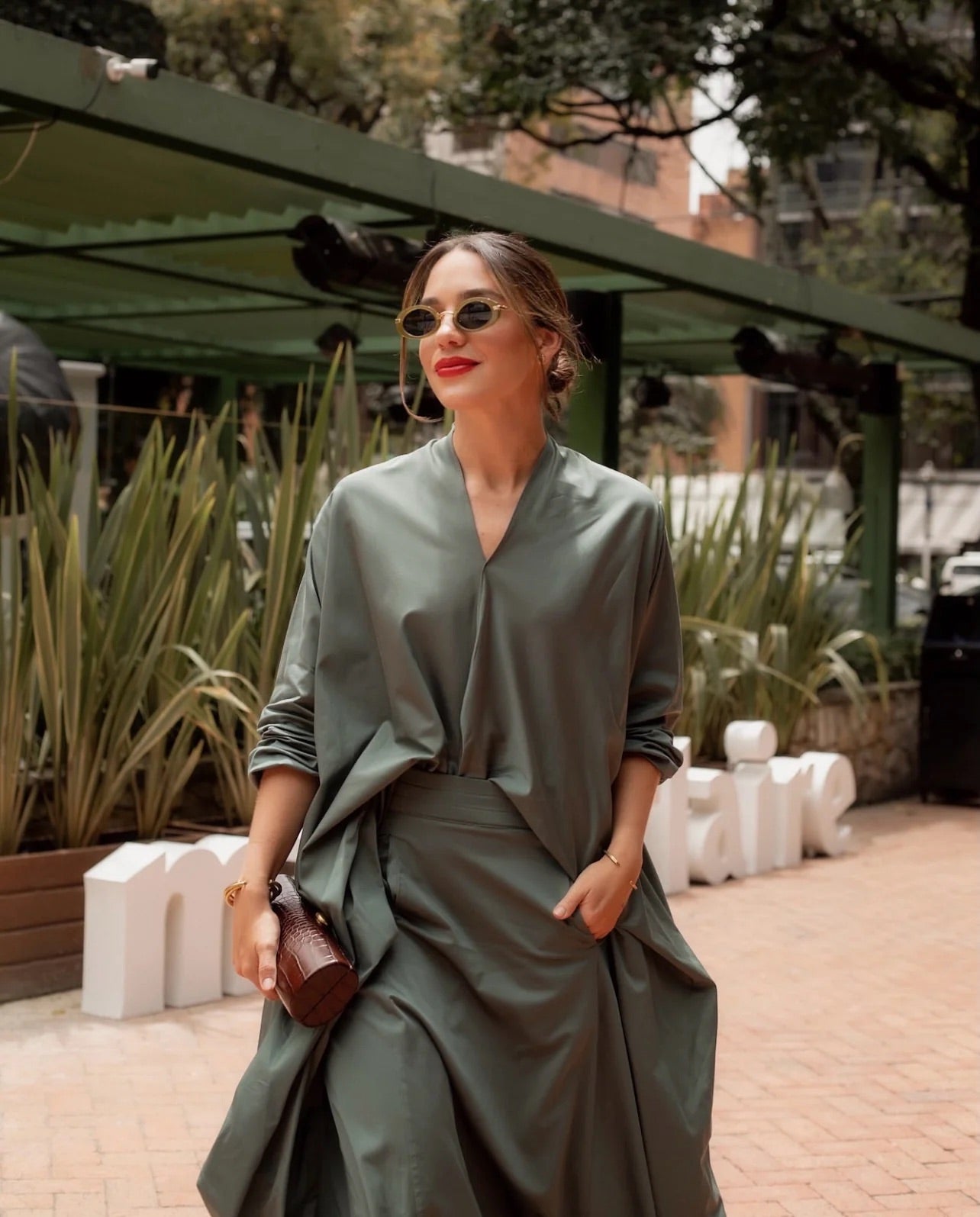 Woman in a green outfit standing outdoors with plants and a building in the background