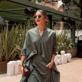 Woman in a green outfit standing outdoors with plants and a building in the background