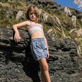 Young girl in a striped top and blue shorts standing on a rocky outcrop with mountains and blue sky in the background.