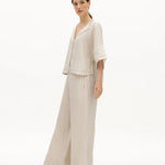 Image of a woman wearing a sand-colored linen top and a matching bottom. The top has a wing collar and the bottom is long and flowy. The purpose of the image is to highlight the clothing by NAOS because it's available for sale at EVAMAIA boutique in Studio City, CA.