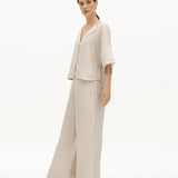 Image of a woman wearing a sand-colored linen top and a matching bottom. The top has a wing collar and the bottom is long and flowy. The purpose of the image is to highlight the clothing by NAOS because it's available for sale at EVAMAIA boutique in Studio City, CA.