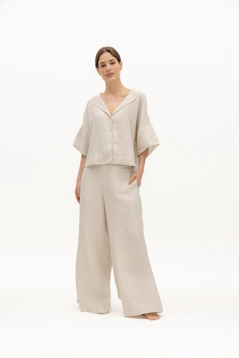 Image of a woman wearing a sand-colored linen top and a matching bottom. The top has a wing collar and the bottom is long and flowy. The purpose of the image is to highlight the clothing by NAOS because it's available for sale at EVAMAIA boutique in Studio City, CA.